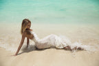 Woman in a white shell dress sitting on a beach with clear blue water.