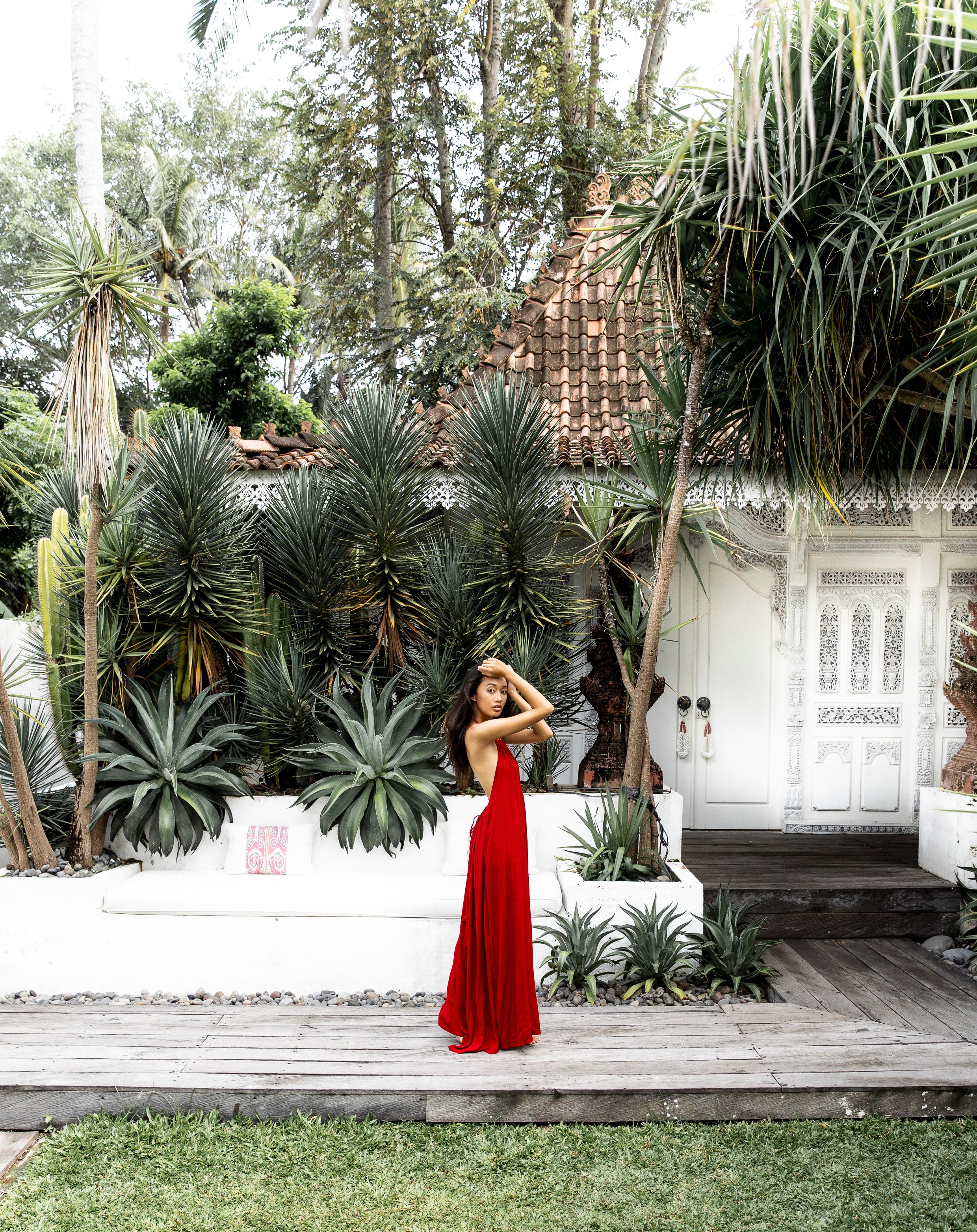 Woman in a red dress standing in a garden with tropical plants and a white Bali cottage