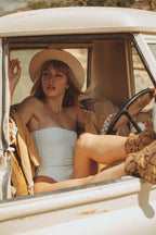 Woman sitting in an open car wearing a white swimsuit and beige hat.