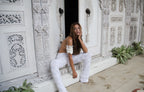 Woman in a white crochet outfit sitting on a decorative white door with plants around.