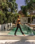 Woman walking by a pool in black crochet beachwear with lush greenery and a building in the background