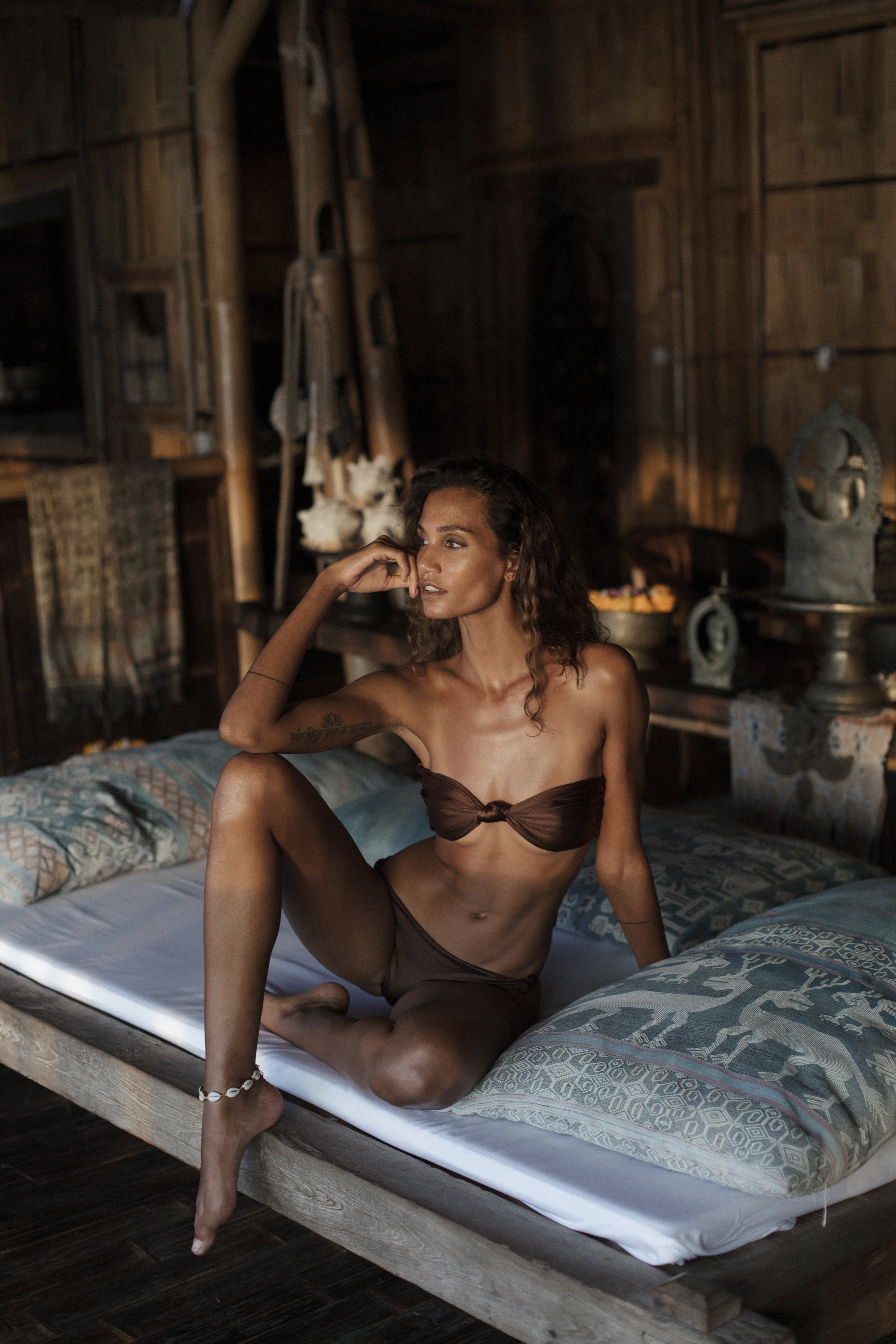 Woman in brown bikini sitting on a bed in a rustic room with wooden walls and various objects.