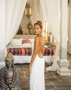 Woman in a white backless dress standing in a bedroom with a Buddha statue and draped curtains.